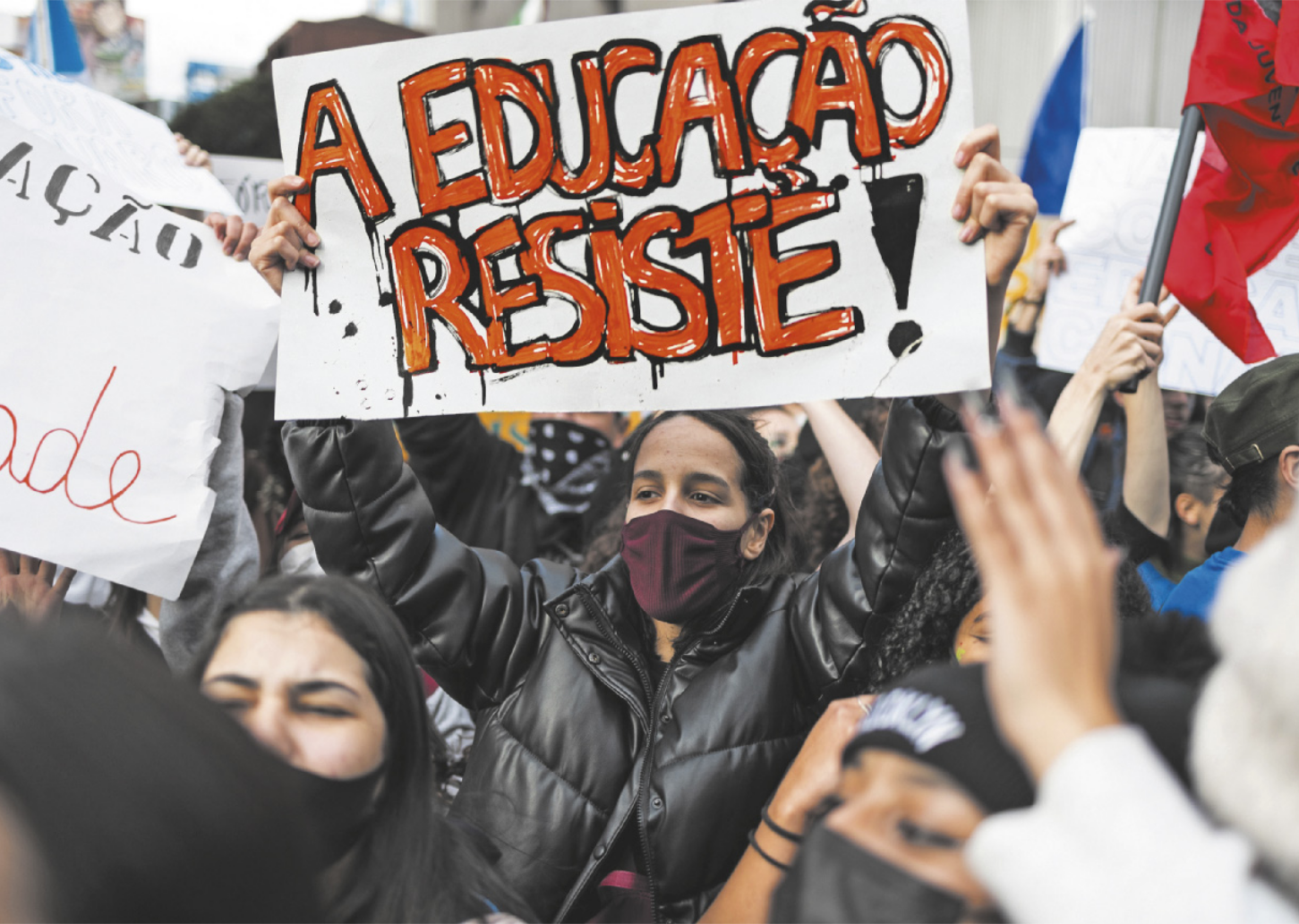 Foto de pessoas em manifestação. Dentre elas, uma jovem leva um cartaz, onde está escrito 'A educação resiste'.
